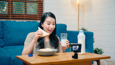 Asian influencer women using camera streaming and recording vlog video about the benefit of drinking milk and sandwiches for breakfast on sofa in living room at home for broadcast online.の写真素材