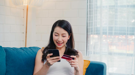 Young smiling Asian woman using smartphone buying online shopping by credit card while lying on sofa when relax in living room at home. Lifestyle women at house concept.の写真素材