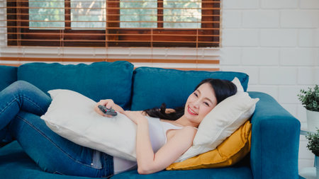 Young Asian woman watching TV at home, female feeling happy lying on sofa in living room. Lifestyle woman relax in morning at home concept.の写真素材