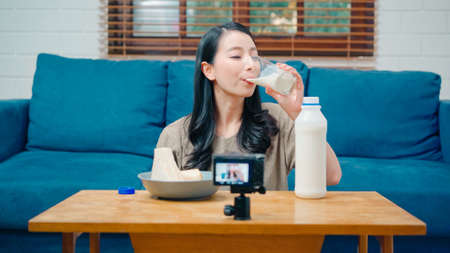 Asian influencer women using camera streaming and recording vlog video about the benefit of drinking milk and sandwiches for breakfast on sofa in living room at home for broadcast online.の写真素材