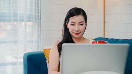 Young business freelance Asian woman working on laptop checking social media and drinking coffee while lying on the sofa when relax in living room at home. Lifestyle women at house concept.の写真素材