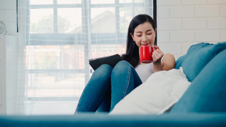 Beautiful attractive smiling Asian woman using tablet holding a warm cup of coffee or tea while lying on the sofa when relax in living room at home. Lifestyle women at home concept.の写真素材