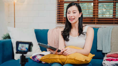Asian fashion influencer designer women using camera streaming and live to sell clothes business, e-commerce broadcast online using tablet chat answer customer on sofa in living room at home.の写真素材
