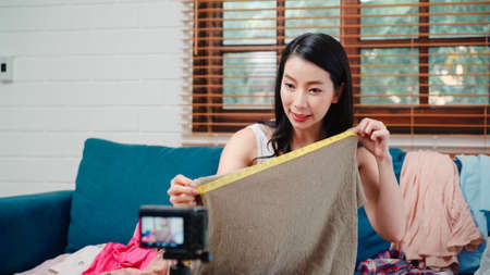 Asian stylist fashion influencer designer women using camera streaming and live to sell clothes, product, and accessories business, e-commerce broadcast online on sofa in living room at home.の写真素材