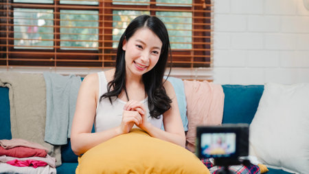 Asian stylist fashion influencer designer women using camera streaming and live to sell clothes, product, and accessories business, e-commerce broadcast online on sofa in living room at home.の写真素材