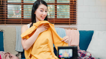 Asian stylist fashion influencer designer women using camera streaming and live to sell clothes, product, and accessories business, e-commerce broadcast online on sofa in living room at home.の写真素材