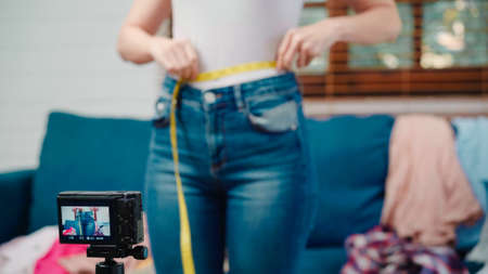 Asian stylist fashion influencer designer women using camera streaming and live to sell clothes, product, and accessories business, e-commerce broadcast online on sofa in living room at home.の写真素材
