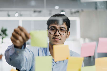 Asian creative man stick a sticky note on glass board. Young professional business male write information, strategy, reminder on paper, business situation, startup in Loft office concept.の写真素材