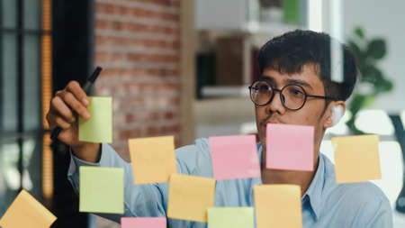 Asian creative man stick a sticky note on glass board. Young professional business male write information, strategy, reminder on paper, business situation, startup in Loft office concept.の写真素材