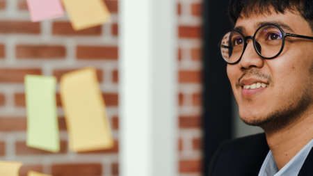 Asian creative man stick a sticky note on glass board. Young professional business male write information, strategy, reminder on paper, business situation, startup in Loft office concept.の写真素材