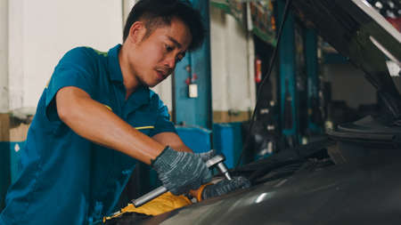 Professional car mechanic screwing details of car engine on lifted automobile at repair service station. Skillful Asian guy in uniform fixing car at mechanics garage at night.の写真素材