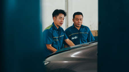 Two professional car mechanic screwing details of car engine on lifted automobile at repair service station. Skillful Asian guy in uniform fixing car at garage night. Car service concept.の写真素材