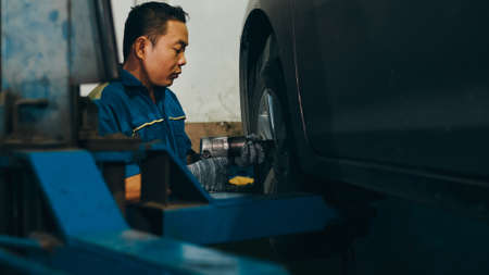 Professional car mechanic screwing details of car engine on lifted automobile at repair service station. Skillful Asian guy in uniform fixing car at mechanics garage at night.の写真素材