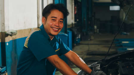 Professional car mechanic screwing details of car engine on lifted automobile at repair service station. Skillful Asian guy in uniform fixing car at mechanics garage at night.の写真素材