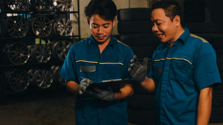 Two professional car mechanic screwing details of car engine on lifted automobile at repair service station. Skillful Asian guy in uniform fixing car at garage night. Car service concept.の写真素材