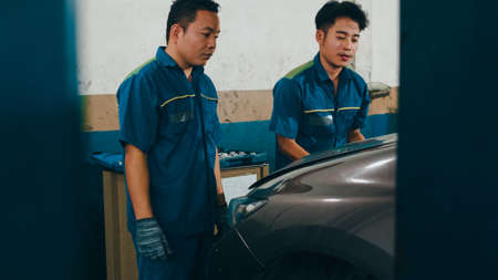 Two professional car mechanic screwing details of car engine on lifted automobile at repair service station. Skillful Asian guy in uniform fixing car at garage night. Car service concept.の写真素材
