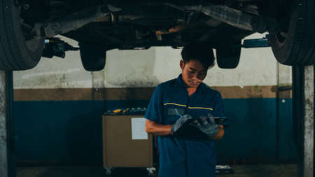 Professional car mechanic screwing details of car engine on lifted automobile at repair service station. Skillful Asian guy in uniform fixing car at mechanics garage at night.の写真素材