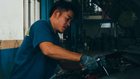 Professional car mechanic screwing details of car engine on lifted automobile at repair service station. Skillful Asian guy in uniform fixing car at mechanics garage at night.の写真素材