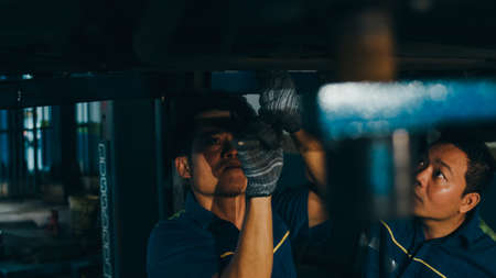 Two professional car mechanic screwing details of car engine on lifted automobile at repair service station. Skillful Asian guy in uniform fixing car at garage night. Car service concept.の写真素材