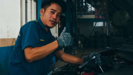 Professional car mechanic screwing details of car engine on lifted automobile at repair service station. Skillful Asian guy in uniform fixing car at mechanics garage at night.の写真素材