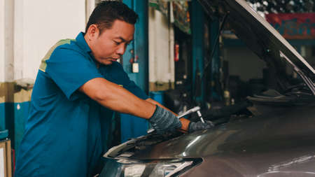 Professional car mechanic screwing details of car engine on lifted automobile at repair service station. Skillful Asian guy in uniform fixing car at mechanics garage at night.の写真素材