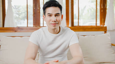 Happy young freelance asia man looking at camera smiling and cheerful relax on video call online on sofa in living room at home, Stay at house quarantine, work from home, Social distancing concept.の写真素材