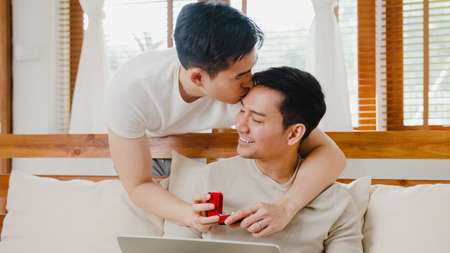 Young Asia gay couple propose at modern home, LGBTQ men happy smiling have romantic time while proposing and marriage surprise wear wedding ring in living room at house. Anniversary romantic concept.の写真素材