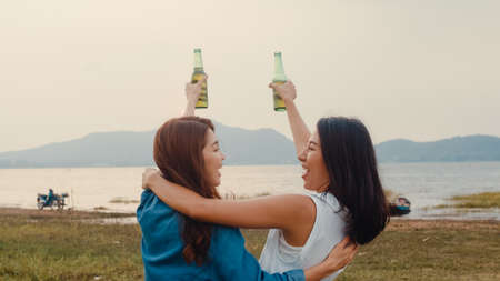 Group of Asia girl couple best friends teenagers drinking have fun salute toast of bottle beer enjoy party with happy moments together in campsite. On background beautiful nature, mountains and lake.の写真素材