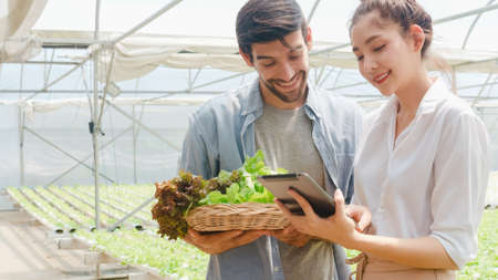 Young Asian businesswoman farmer hold tablet show performance of her organic farm to hispanic man buyer at greenhouse farm garden, New innovation technology in the cultivation concept.の写真素材