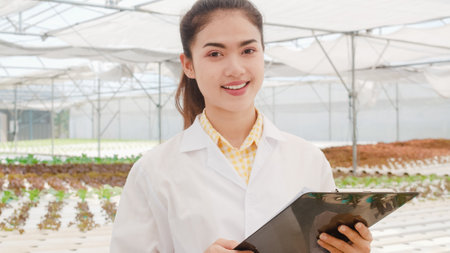 Close-up Happy Asian scientist woman looking at camera with smile hold document for research and development in organic vegetable at greenhouse farm garden, New technology in the cultivation concept.の写真素材