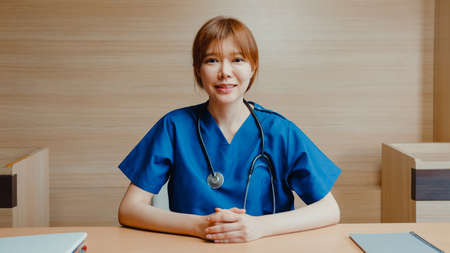 Young Asia lady doctor in white medical uniform with stethoscope using computer laptop talk video conference call with patient, looking at camera in health hospital. Consulting and therapy concept.の写真素材