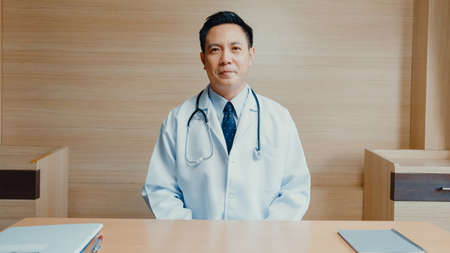 Young Asia guy doctor in white medical uniform with stethoscope using computer laptop talking video conference call with patient at desk in health clinic or hospital. Consulting and therapy concept.の写真素材