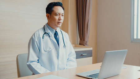 Young Asia guy doctor in white medical uniform with stethoscope using computer laptop talking video conference call with patient at desk in health clinic or hospital. Consulting and therapy concept.の写真素材