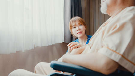 Caring young Asia nurse lady support assist positive old guy patient sit on wheelchair at home or hospital. Rehabilitation activity, elderly healthcare promotion, healthcare and medicine concept.の写真素材
