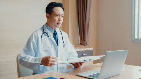 Young Asia guy doctor in white medical uniform with stethoscope using computer laptop talking video conference call with patient at desk in health clinic or hospital. Consulting and therapy concept.の写真素材