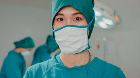 Young Asian nurse woman looking at camera and smiling after performing invasive surgery on patient in hospital operating room. Diverse team of professional surgeon, Healthcare and medical concept.の写真素材