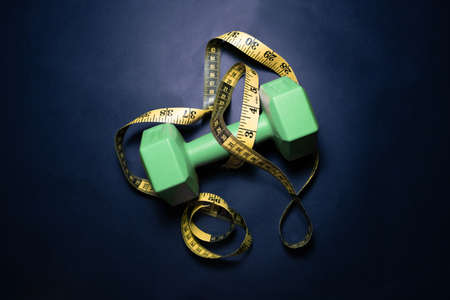 Dark sports concept. Sports equipment as dumbbells, expander with pink handles and centimeter tape on a dark black cement background, top view with a copy space.の写真素材