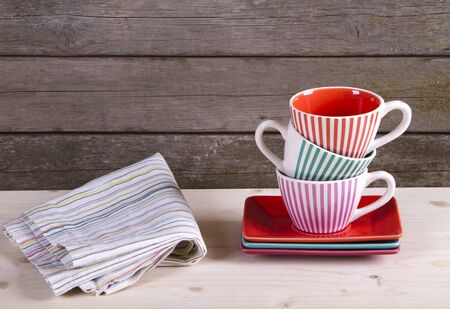 Colorful striped coffee cups on shelf and cotton towel against rustic old vintage wooden wall with copy spaceの写真素材