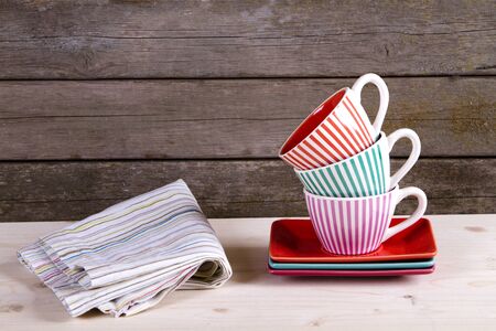 Colorful striped coffee cups on shelf and cotton towel against rustic old vintage wooden wall with copy spaceの写真素材