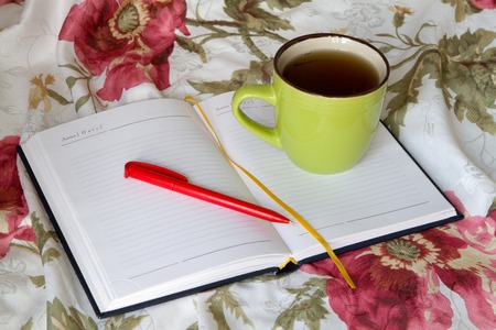 Green mug with tea or coffee in bed with bedding with floral print diary with a pencil or pen note thoughts dreamsの写真素材