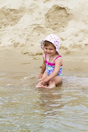 Little cute girl standing in the sea water on the beach in a swimsuit and a hat in summer on vacationの写真素材