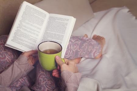 Woman sitting on sofa, reading book and drinkの写真素材