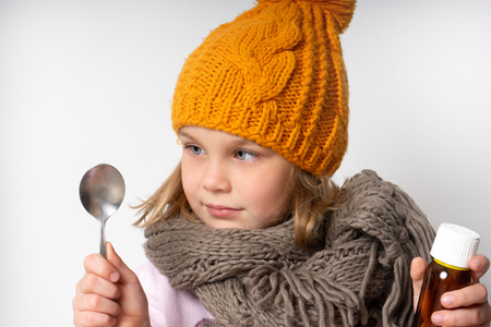 Little woolen dress in a warm scarf and knitted hat. holds in his hands a bottle of cough syrup for colds and looks at the spoon, is going to drink.の写真素材