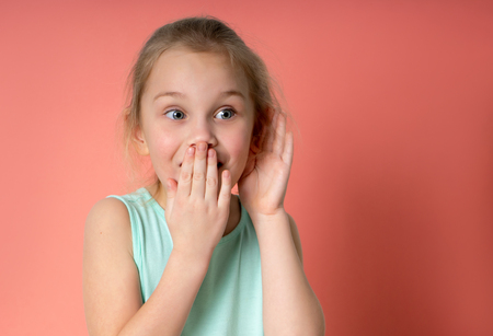 Colorful studio portrait of pretty little child girl in blue dress with palm near her ear. She covers her mouth with her hand in surprise. Gossip concept.の写真素材