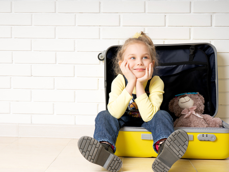 Little cute girl in denim overalls and boots. sits with his friend a teddy bear in an opened suitcase. Ready to travel. we pack thingsの写真素材
