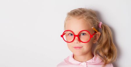 Little blonde child with ponytail, in red framed glasses, pink blouse. She is smiling, posing with yellow pencil behind her ear. Isolated on white. Childhood, online education. Close up, copy space