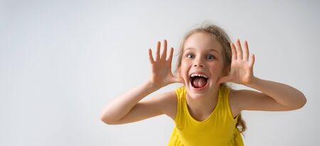 Sly happy preschool girl in sunny yellow dress teasing someone, yelling and laughing with funny face. Happy childhood, emotions, gesturing, playing, having fun. Close up studio shot isolated on whiteの写真素材