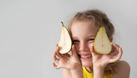 Cute little blond girl with cheerful smile peeking out from behind two halves of pear she is holding in hands. Happy childhood, facial expression, games and fun. Close up shot isolated on whiteの写真素材