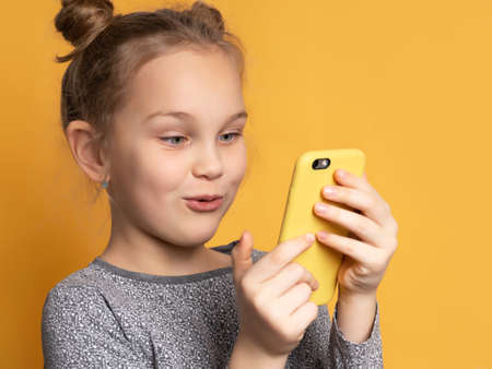 Attractive little blonde girl looking at her mobile phone screen with joyful face, on yellow background for copy spaceの写真素材