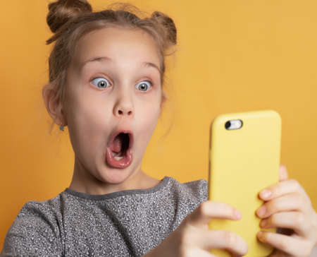 Close up of surprised little girl with wide open mouth looking at phone holding in hands. Portrait of a shocked child on a yellow background. Technology, active lifestyle conceptの写真素材